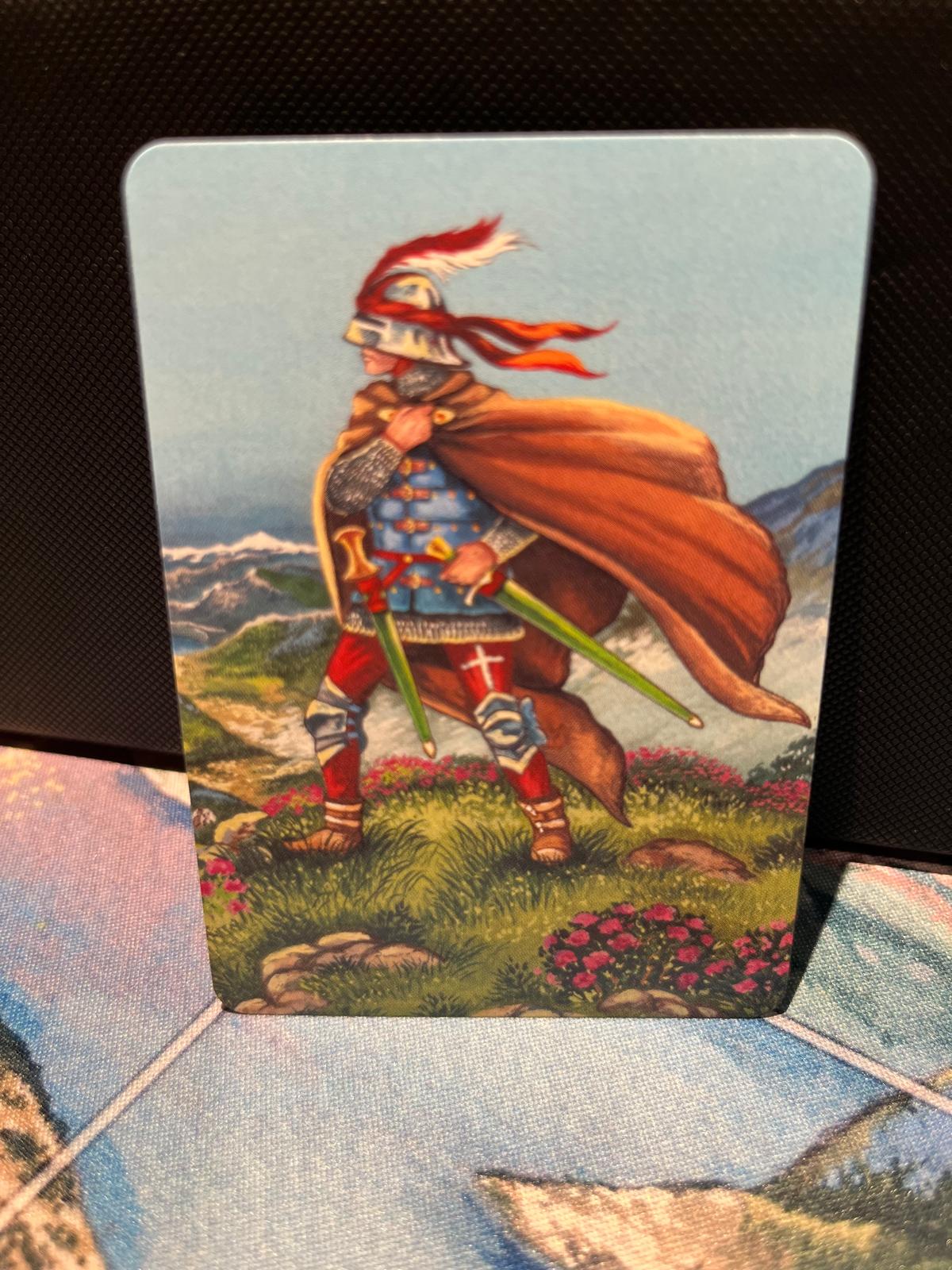 Foot Soldier promo token — photograph of the physical “Foot Soldier” trading card standing on a patterned playmat, showing a knight with a red and white plume, green swords, and pink flowers before snow-capped mountains
